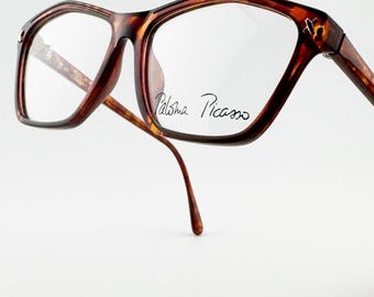 May include: Brown tortoiseshell eyeglasses with a rectangular frame. The glasses have clear lenses and the text "Paloma Picasso" is written on the lens. The frame has a unique shape and design.
