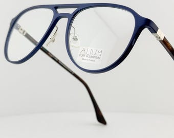 May include: Blue aviator-style eyeglasses with clear lenses. The frames are a deep blue, with a bridge connecting the lenses. The temples are a dark brown with a silver accent. The text "ALIUM PURE ALUMINIUM Made in France" is visible on the lens.