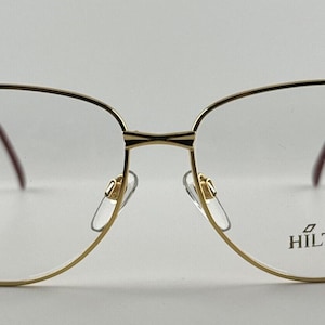 May include: Gold-tone metal eyeglasses with a delicate chain detail on the frame. The glasses have a clear lens and the brand name "HILTON" is printed on the right lens.