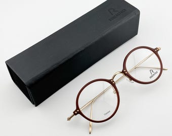 May include: Round eyeglasses with a burgundy frame and gold-colored arms, next to a black rectangular case. The glasses have the word "Titanium" printed on the lens and the brand name "RODENSTOCK" on the case and lens.