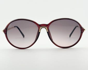 May include: Round-framed sunglasses with a burgundy frame and gradient lenses. The bridge and temples feature gold-colored accents. The lenses transition from a darker shade at the top to a lighter shade at the bottom.