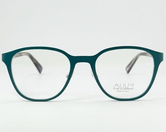 May include: Teal-colored eyeglasses with a round frame. The glasses have clear lenses and the text "ALIUM PURE ALUMINIUM Made in France" is printed on the right lens. The frames are a solid teal color.