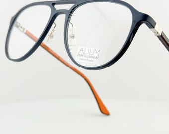 May include: Aviator-style eyeglasses with a navy blue frame and clear lenses. The temples are brown with orange tips. The glasses have a silver bridge and the text "ALIUM PURE ALUMINIUM Made in France" is visible on the lens.