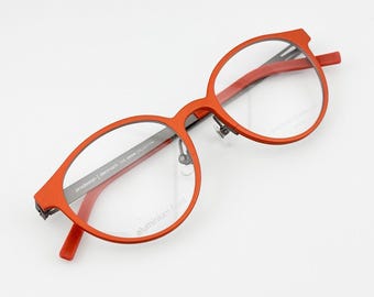 May include: Red-framed eyeglasses with a round shape and clear lenses. The glasses have a modern design with a thin frame and a bridge. The text "prodesign denmark THE AXION COLLECTION" is printed on the arm. The text "aluminium front" is printed on the lens.