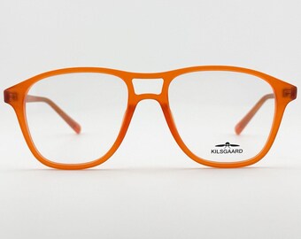 May include: Orange-framed eyeglasses with a classic aviator shape. The frames are a solid, vibrant orange, with a bridge connecting the two lenses. The temples are a lighter shade of orange. The brand name "KILSGAARD" is printed on the lower right lens.