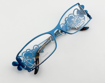 May include: Blue eyeglasses with a floral design on the frames. The lenses are clear, and the frames have a decorative cutout pattern. The text "Icons" is visible on the lens. The glasses are made of titanium.