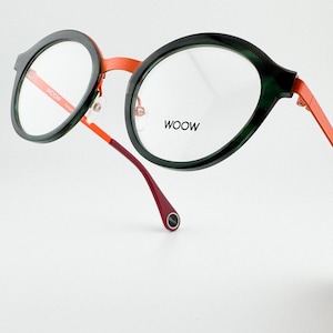 Może przedstawiać: Okulary okrągłe w zielono-czarnej oprawie z pomarańczowymi i czerwonymi zausznikami. Okulary mają napis "WOOOW" nadrukowany na prawej soczewce.