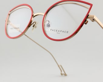 May include: Gold-toned eyeglasses with red rims. The frames feature a unique, angular design. The lenses are clear, and the text "FACE A FACE PARIS" is visible on one lens. The text "PURE TITANIUM" is visible on the other lens.