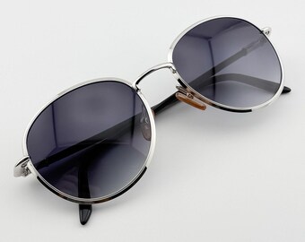 May include: Silver-framed sunglasses with dark gray lenses. The round lenses are set in a thin, silver metal frame. The temples are black with a small, light brown piece at the hinge. The sunglasses are set against a white background.