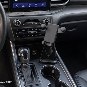 May include: A black adjustable smartphone mount installed in the center console of a 2022 Ford Explorer. The mount features a rectangular cradle with side arms and a base that sits near the gear shifter and cup holders, with the text Ford Explorer 2022.
