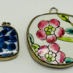 May include: Two pendants with metal frames. The smaller pendant features a blue and white floral pattern. The larger, circular pendant displays a white background with pink flowers, green leaves, and a yellow center, all encased in a metallic border.