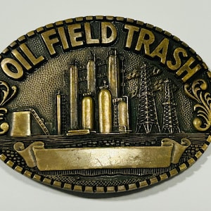 May include: An oval-shaped brass-toned metal belt buckle featuring a raised relief design of an oil refinery with towers and derricks. The text OIL FIELD TRASH is embossed along the top edge, and a blank banner is centered at the bottom.