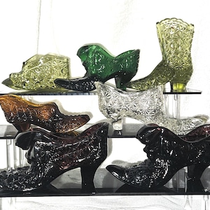 May include: A collection of vintage glass shoe figurines in various colors, including green, amber, clear, and black. The shoes are arranged on tiered shelves, showcasing their intricate starburst patterns and high heels. The collection includes a boot and several heeled shoe styles.