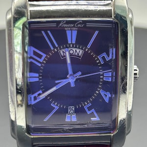 May include: A rectangular Kenneth Cole New York wristwatch with a dark blue face and silver-tone case. The watch features blue numerals, hands, and a date display. The strap is a reddish-brown leather.