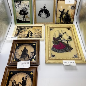 May include: A collection of framed silhouette art pieces. The pieces depict various scenes, including figures in period clothing, couples, and a woman at a spinning wheel. The frames are in various colors and styles, including green, gold, and brown.