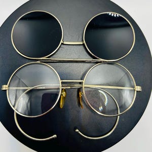May include: Gold-rimmed round sunglasses with black lenses above clear-lensed glasses. The glasses have a double-bridge design and curved metal accents. The lenses are set against a black background.