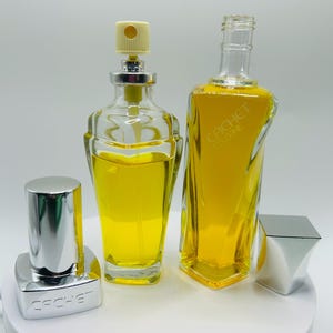 May include: Two glass perfume bottles filled with yellow liquid. One bottle has a silver cap with the word CACHET embossed on it, while the other is a tall rectangular bottle with CACHET COLOGNE etched on the front and a separate silver cap.