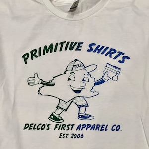 May include: White t-shirt with a green and blue graphic of a cartoon character wearing a hat that says "Delco" and holding a paintbrush. The text "Primitive Shirts" is above the character and "Delco's First Apparel Co. Est. 2006" is below the character.