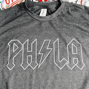 May include: A dark gray t-shirt with the white text "PHLA" in a bold, angular font, with a lightning bolt replacing the "I". The shirt has a crew neck and is made of a soft material. The background has various stickers.