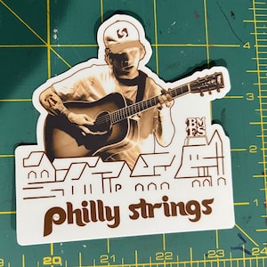 May include: A white sticker with a brown silhouette of a person playing a guitar. The sticker has the text "philly strings" in brown letters and a small building with the letters "BMFS" on top.