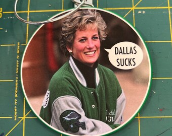 Op de afbeelding: Ronde kerstversiering met een lachende vrouw die een groene en grijze varsity-jas draagt. De versiering heeft de tekst "DALLAS SUCKS" in een tekstballon. Het heeft een zilveren metalen lus om op te hangen.