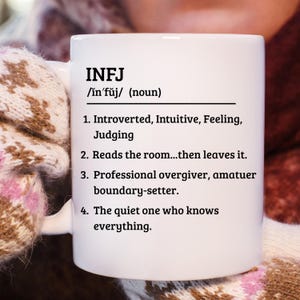 May include: White ceramic mug with the text "INFJ" and a definition. The definition includes the pronunciation, a list of personality traits, and a description of the INFJ personality type. The mug is held by a person wearing a patterned glove.