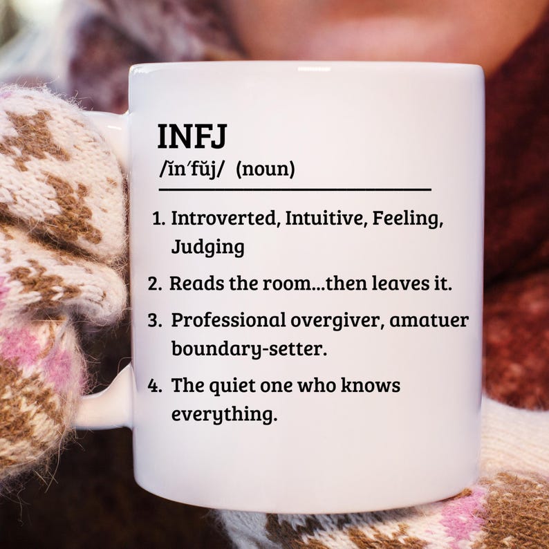 May include: White ceramic mug with the text "INFJ" and a definition. The definition includes the pronunciation, a list of personality traits, and a description of the INFJ personality type. The mug is held by a person wearing a patterned glove.