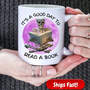 Puede incluir: Taza de cerámica blanca con el texto "IT'S A GOOD DAY TO READ A BOOK" alrededor de un gráfico de luna púrpura. El gráfico presenta un esqueleto leyendo un libro. La taza tiene un asa blanca.