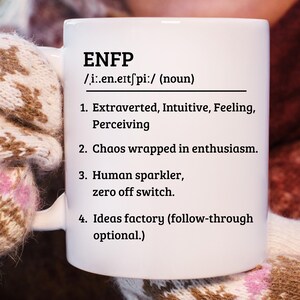 May include: White ceramic mug with the text "ENFP" and a definition including "Extraverted, Intuitive, Feeling, Perceiving" and "Chaos wrapped in enthusiasm." The mug is held by a person wearing a patterned glove.