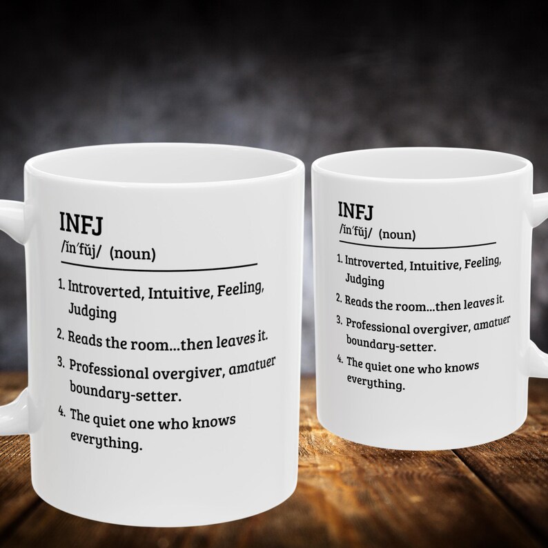 May include: Two white ceramic mugs with the text "INFJ" and a definition printed in black. The mugs are on a wooden surface. The text includes the definition of INFJ, a personality type.