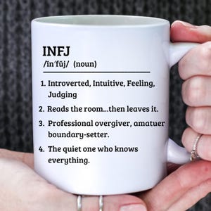 May include: White ceramic mug with the text "INFJ" and a definition, including personality traits and characteristics. The mug is held by a person.