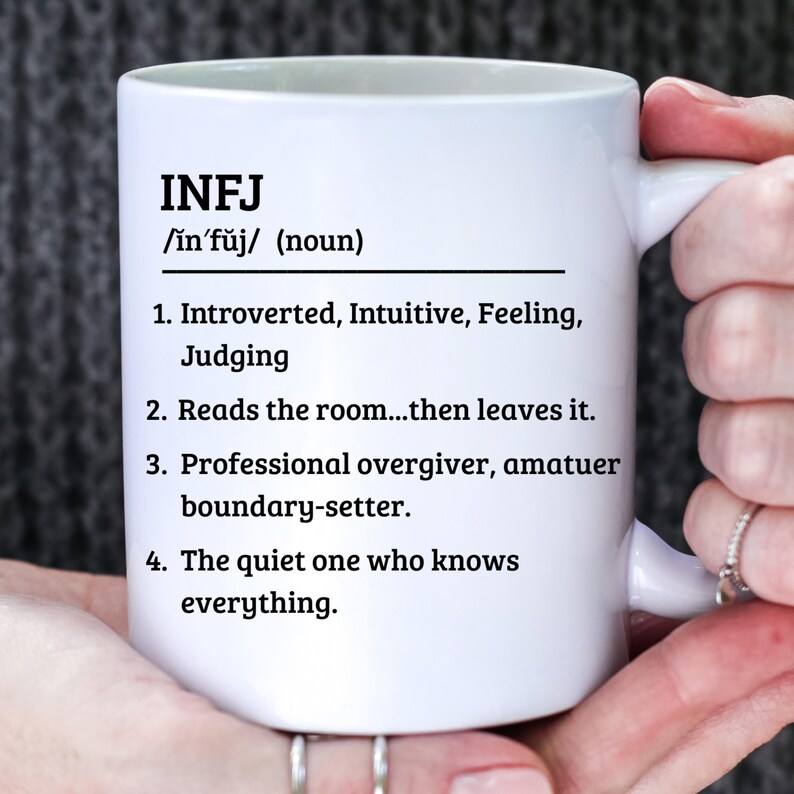 May include: White ceramic mug with the text "INFJ" and a definition, including personality traits and characteristics. The mug is held by a person.