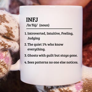 May include: White ceramic mug with the text "INFJ" and a definition. The definition includes the pronunciation, and four descriptive statements about the personality type. The mug is held by a person wearing a patterned glove.