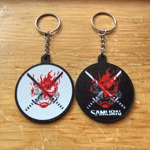May include: Two circular keychains with metal rings and chains. One keychain has a white background with a red and black oni mask graphic and crossed swords. The other has a black background with the same graphic and the text SAMURAI in white.