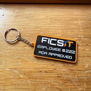 May include: A rectangular keychain with a black textured surface and an orange border. The text reads FICSIT, EMPLOYEE #222, and ADA APPROVED in white lettering. A silver metal split ring is attached to the keychain by a short metal chain.