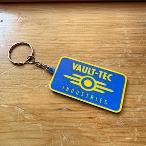 May include: A rectangular keychain featuring a blue background with a yellow border. The text VAULT-TEC INDUSTRIES is printed in yellow, accompanied by a yellow circular logo with horizontal lines extending from the center on both sides.