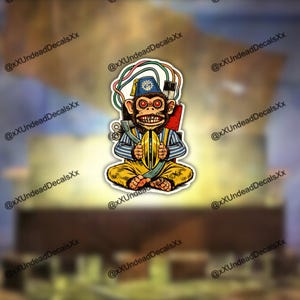 Puede incluir: Una pegatina con un mono de dibujos animados con un fez y sosteniendo un objeto amarillo. El mono tiene ojos rojos, una amplia sonrisa y viste ropa a rayas azules y blancas y pantalones amarillos. Detrás del mono hay cables y una pequeña mochila roja.