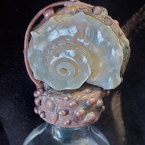 May include: A decorative bottle stopper featuring a pearlescent seashell set in a copper-toned metal base. The stopper is mounted on a clear glass bottle, creating a unique and elegant design. The seashell has a spiral shape.