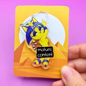 Puede incluir: Un pin de personaje de gato de dibujos animados amarillo y azul con el texto "mature content" en una tarjeta amarilla con un diseño de pirámide y sol. El pin está sujeto por una mano sobre un fondo púrpura.