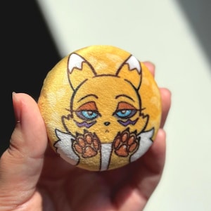 Könnte beinhalten: Ein runder, gelber Knopf mit einer Cartoon-Fuchs-Illustration. Der Fuchs hat blaue Augen, lila Akzente und orange Pfoten. Der Knopf wird in einer Hand gehalten.