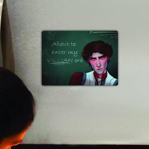 May include: A green chalkboard with white writing that says "About to enter my VILLIAN era". A young man with dark hair and a serious expression is standing in front of the chalkboard. He is wearing a white shirt with a red vest and a black bow tie.