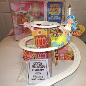 May include: A vintage Candy Mountain Flatsy playset. The set includes a colorful, multi-level structure with a track, a doll, and a yellow vehicle. The box features a child playing with the toy. The set includes a booklet titled "Playing Instructions".