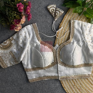 May include: A white blouse with gold embroidery and embellishments. The blouse features a square neckline and short sleeves. The edges are trimmed with gold sequins. The sleeves have intricate gold patterns. The blouse is likely made of silk or a similar fabric.