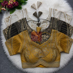 May include: Three short-sleeved blouses in mustard yellow, black, and white, each featuring intricate gold embroidery, a scoop neckline, and a decorative tie-up back with dangling tassels. The garments are layered, showcasing detailed stitching.