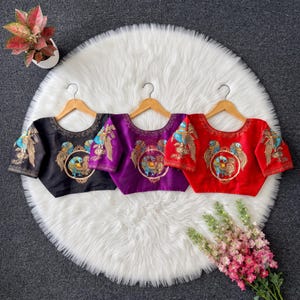 Puede incluir: Tres blusas de manga corta en negro, morado y rojo, cada una con un bordado circular central de un pavo real con hilos dorados, azules y verdes. Las prendas tienen cuello redondo y se muestran colgadas en perchas de madera.