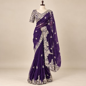 May include: A deep purple saree featuring intricate silver embroidery along the borders and across the bodice. The garment is displayed on a mannequin, showcasing a sheer fabric with floral patterns and a scalloped edge detail throughout.