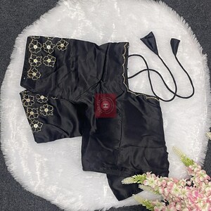 May include: A black satin blouse featuring short sleeves with gold floral embroidery and central sequins. The garment includes thin black tie-up straps at the neckline and a square-cut back design. The fabric has a smooth, reflective finish.
