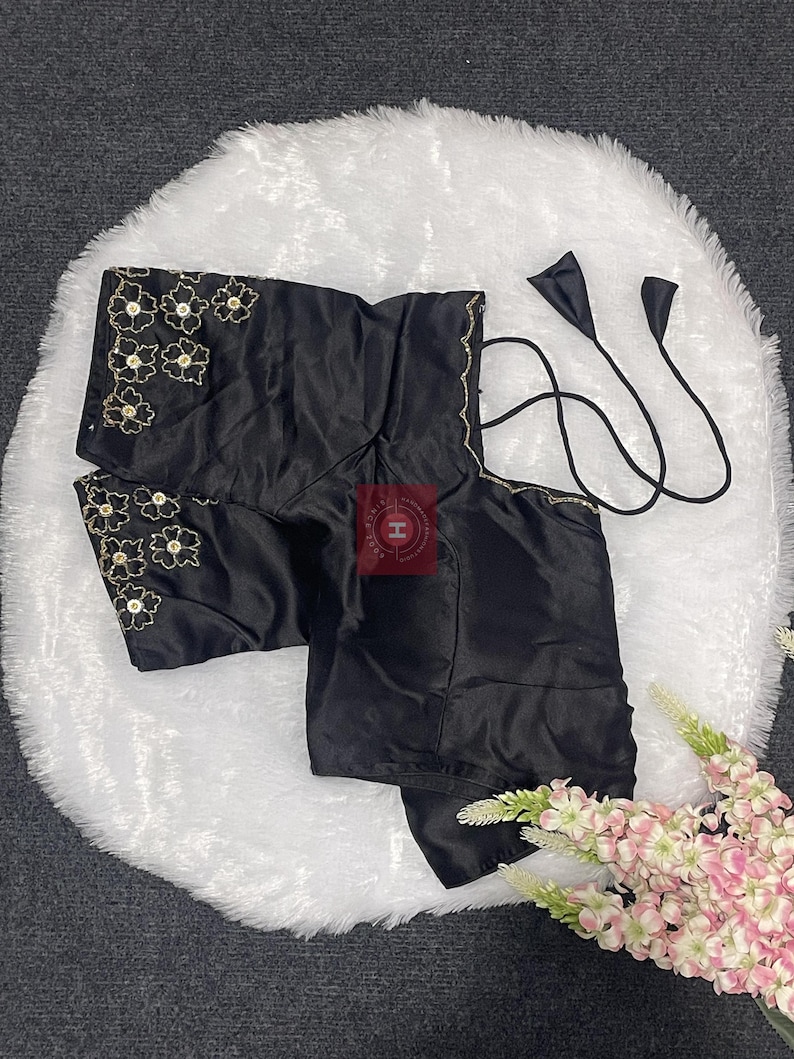 May include: A black satin blouse featuring short sleeves with gold floral embroidery and central sequins. The garment includes thin black tie-up straps at the neckline and a square-cut back design. The fabric has a smooth, reflective finish.