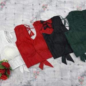 Puede incluir: Una colección de blusas en varios colores: blanco, rojo, negro y verde. Las blusas tienen diferentes estilos, incluyendo opciones sin mangas y de manga corta, con detalles de lazo y espalda abierta. Las blusas están dispuestas sobre una tela estampada.