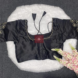 May include: A black blouse with short sleeves featuring gold floral embroidery on the cuffs and neckline. The garment has a square neckline and thin black tie strings at the back. The fabric has a smooth, satin-like finish with a subtle sheen.